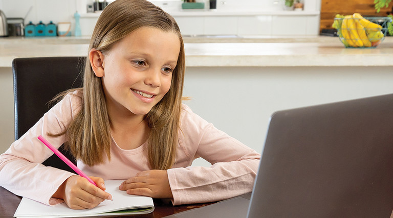 girl smiling and taking notes on paper while looking at computer screen